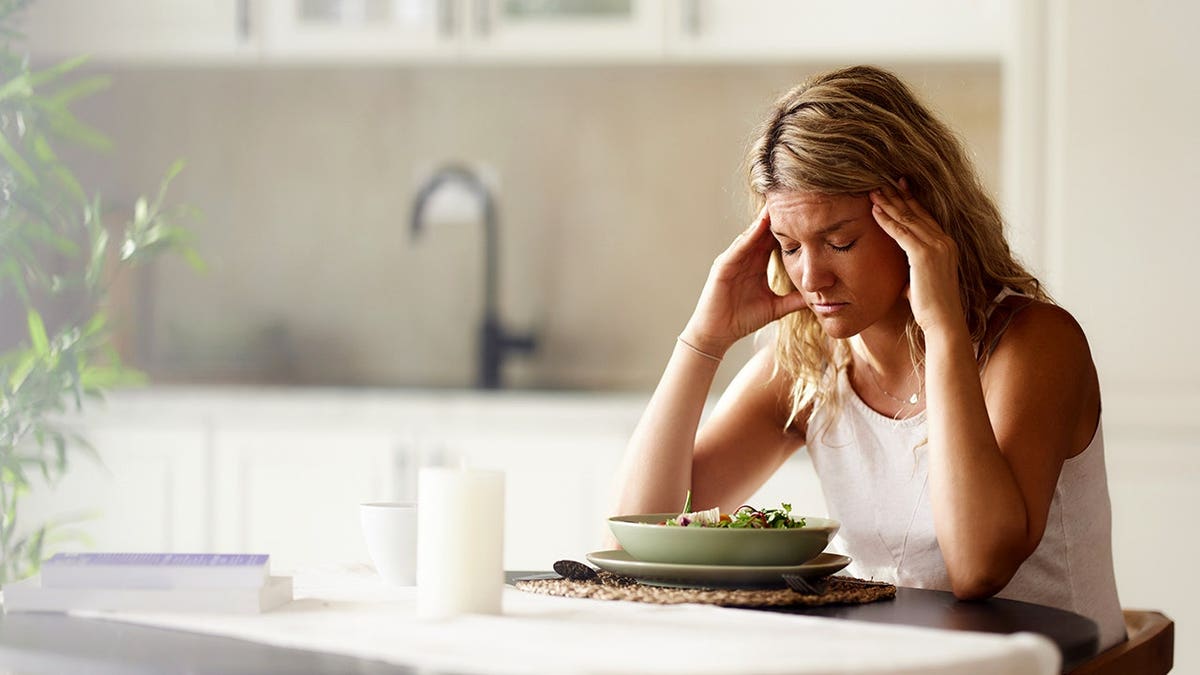 Woman holds her head in pain while seated at a dining table during breakfast.