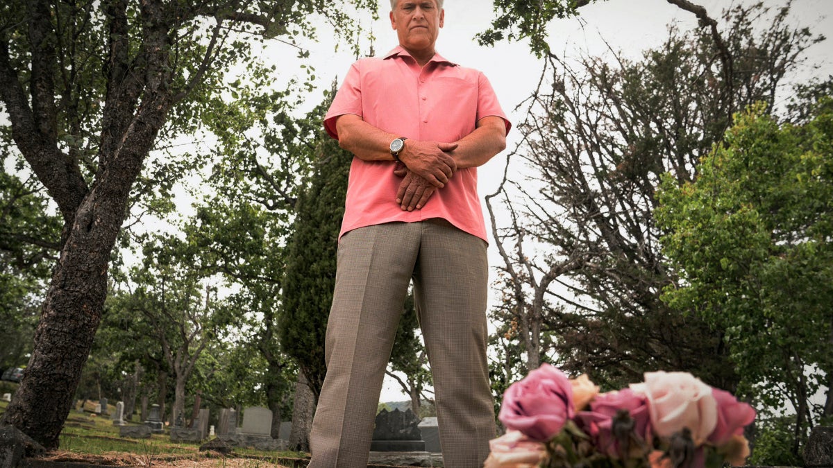 Bruce Campbell stands outdoors in a cemetery while dressed in a pink shirt.