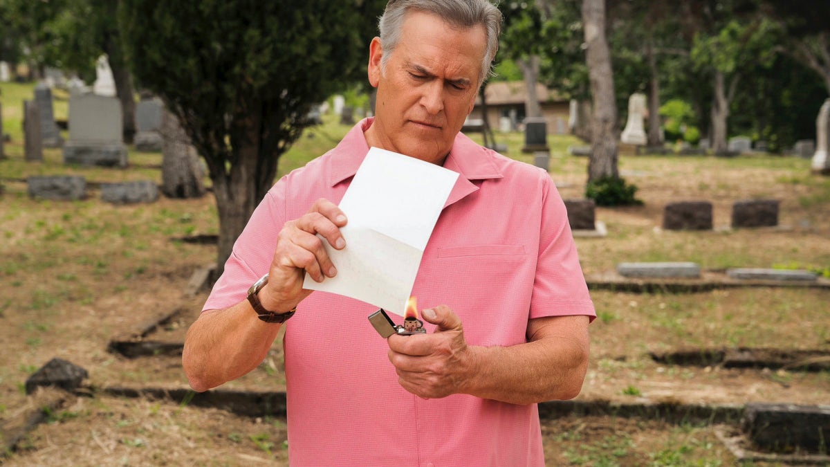 Bruce Campbell uses a lighter to set fire to a piece of paper in a cemetery.