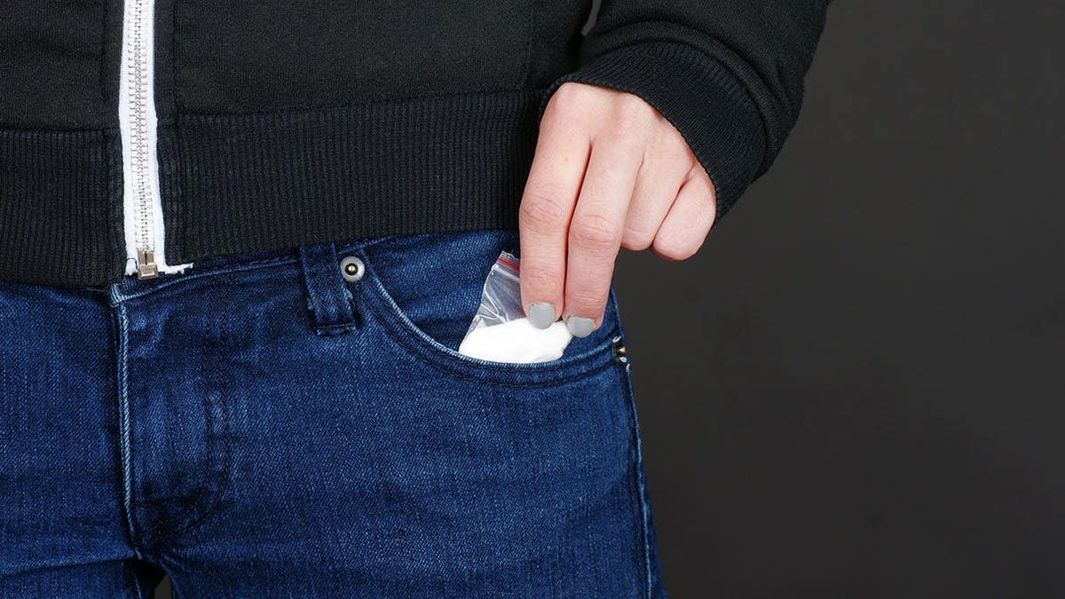 Female hand retrieving drug packet from blue jean pocket against black background.