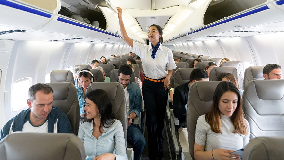 Flight attendant walks down airplane aisle closing overhead compartments and smiling.