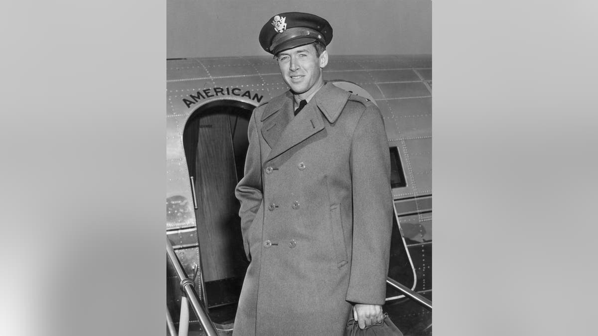Jimmy Stewart in Air Force cap and wool overcoat standing in front of military airplane.