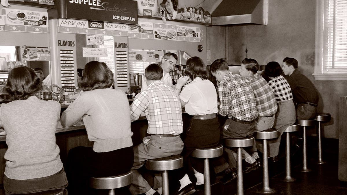 A group of young people sits at a luncheonette in the 1950s.