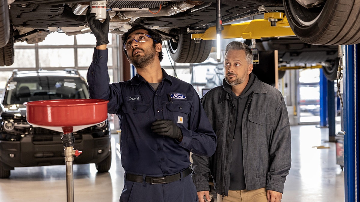 Nate Bargatze looks on as a Ford mechanic works beneath a car.