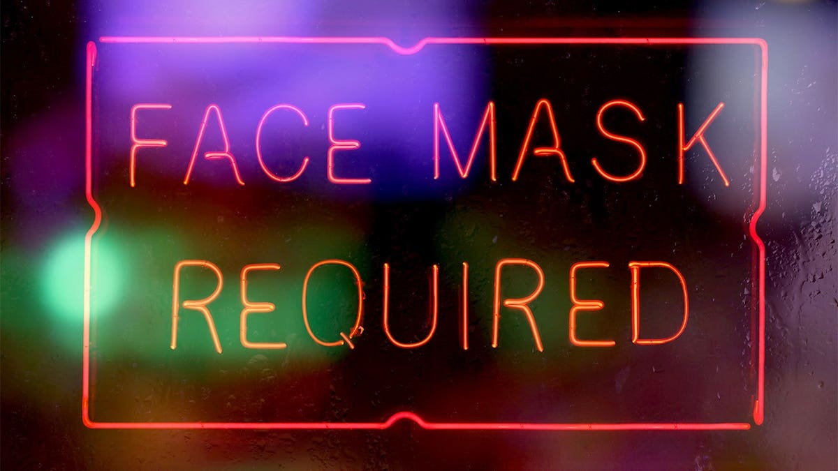 Neon face mask required sign displayed in a rainy window