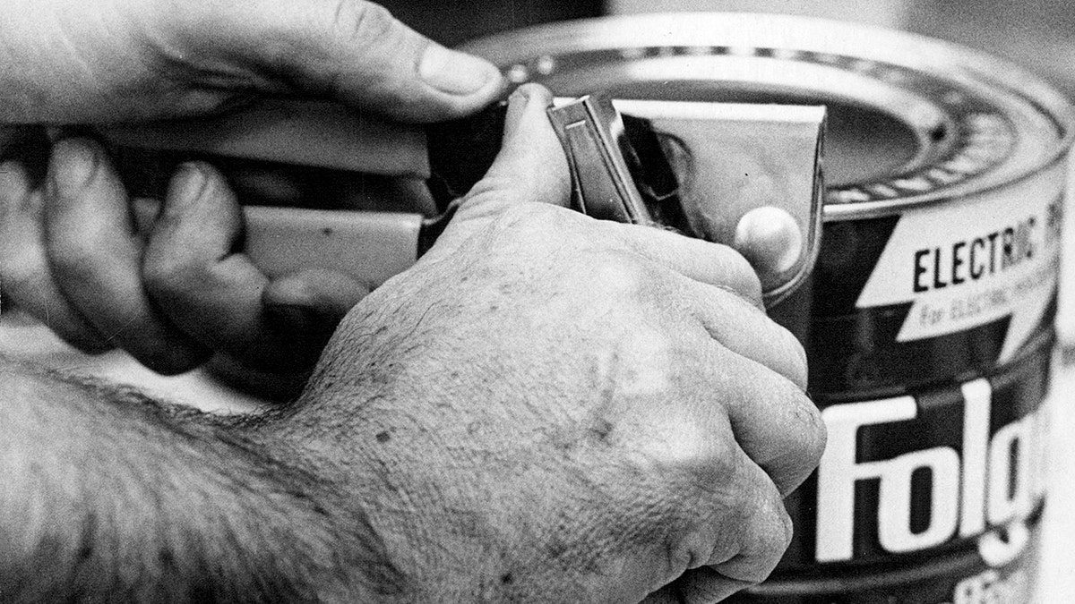 A black and white photo shows a man's hands using a can opener to open a Folgers coffee can.