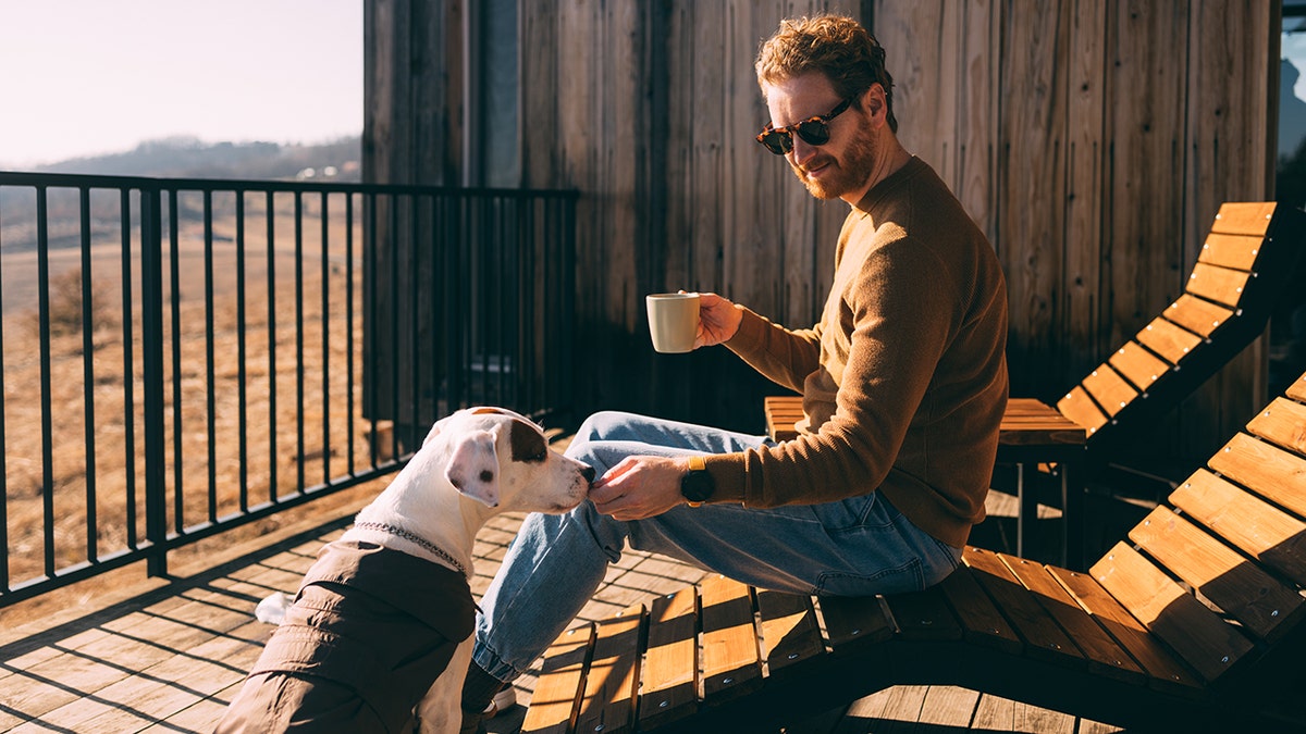 A man holding a coffee cup and wearing sunglasses gives a dog a treat from the patio.