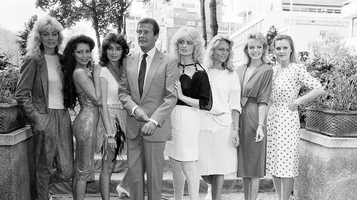 Roger Moore stands with seven women from the James Bond film Octopussy at a press event.