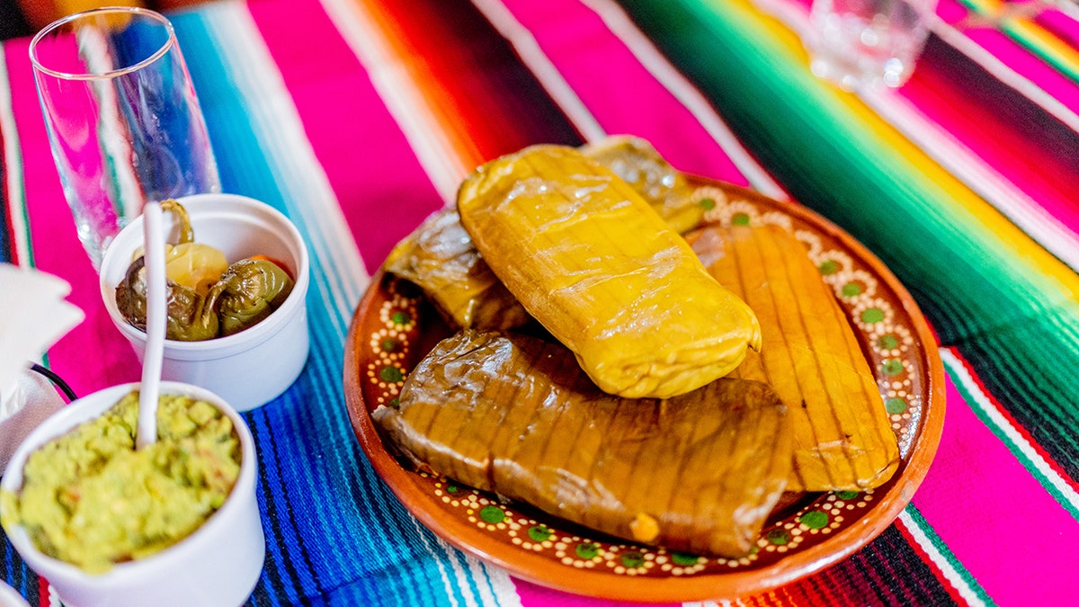A tamale is shown on a plate with other Mexican foods at a table with a colorful cloth.