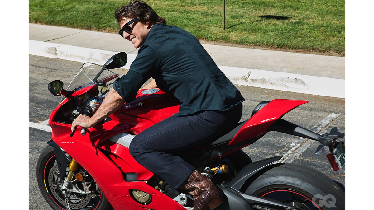 Tom Cruise sits on a red Ducati motorcycle outdoors while wearing sunglasses.