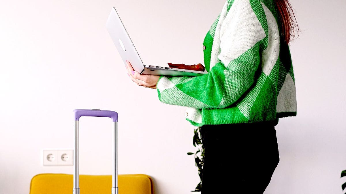 A woman uses a laptop next to a suitcase.