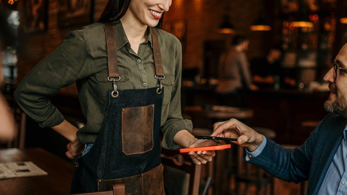 Two businessmen talk at a bar while a waitress swipes a card for payment.