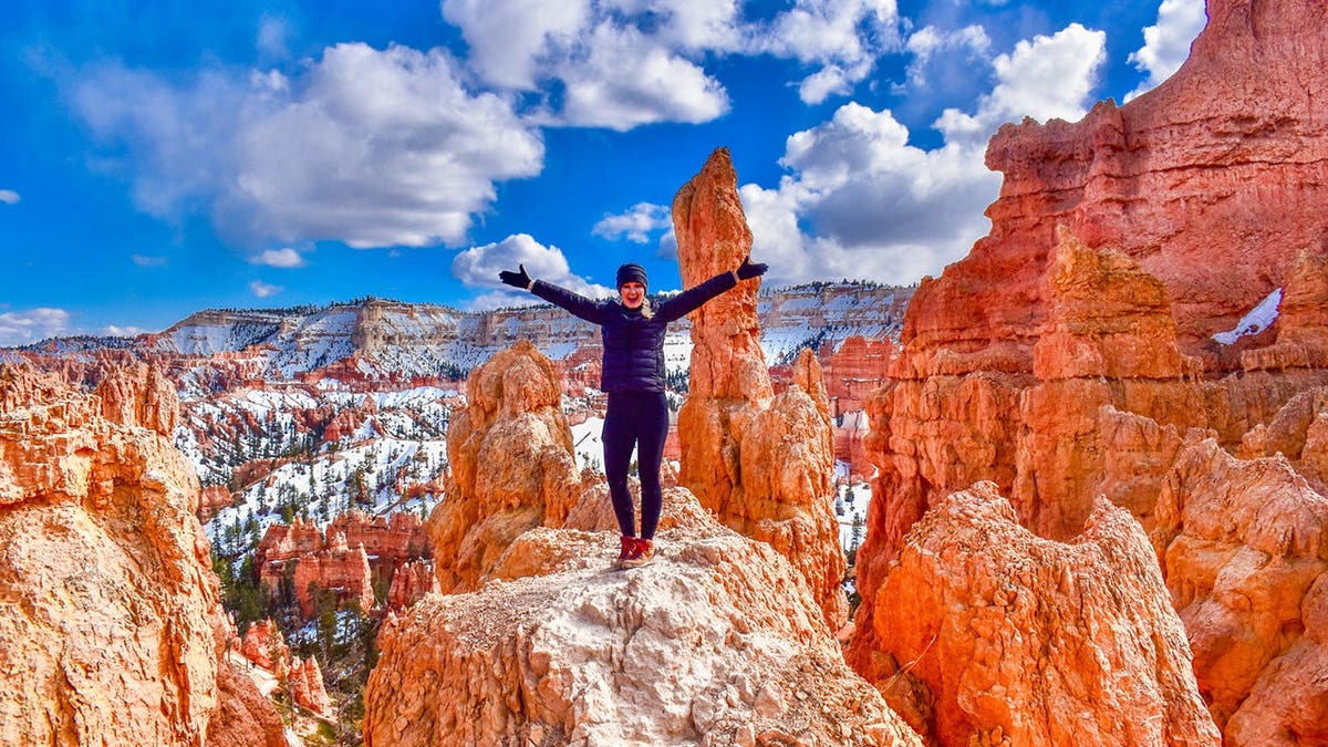 Blonde woman stands in Bryce Canyon National Park with red rock formations and blue sky.