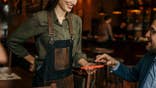Two businessmen talk at a bar while a waitress swipes a card for payment.
