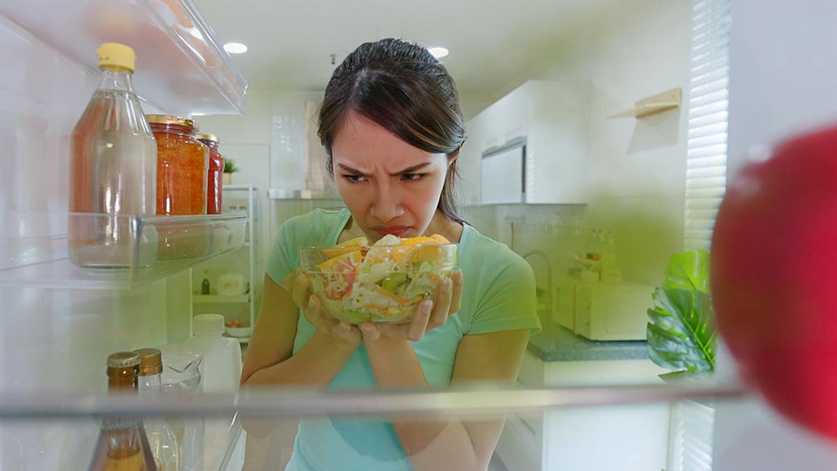 Asian woman opens refrigerator in kitchen, reacting to bad smell from spoiled salad.