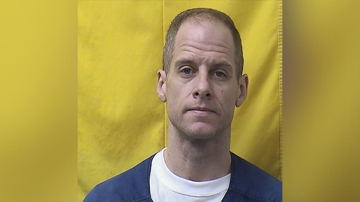 New prison mugshot of convicted wife killer Caleb Flynn wearing a blue prison uniform over a white shirt against a yellow background.