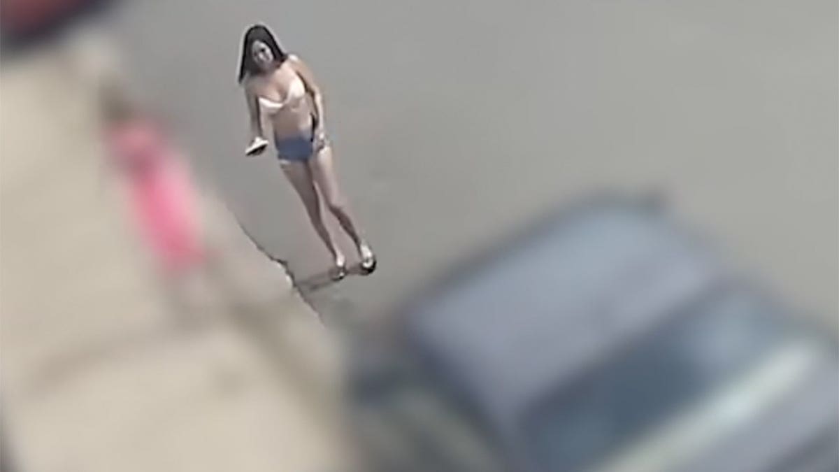 A woman in a light-colored top, denim shorts and sandals faces the camera on a paved street, holding an object in one hand.