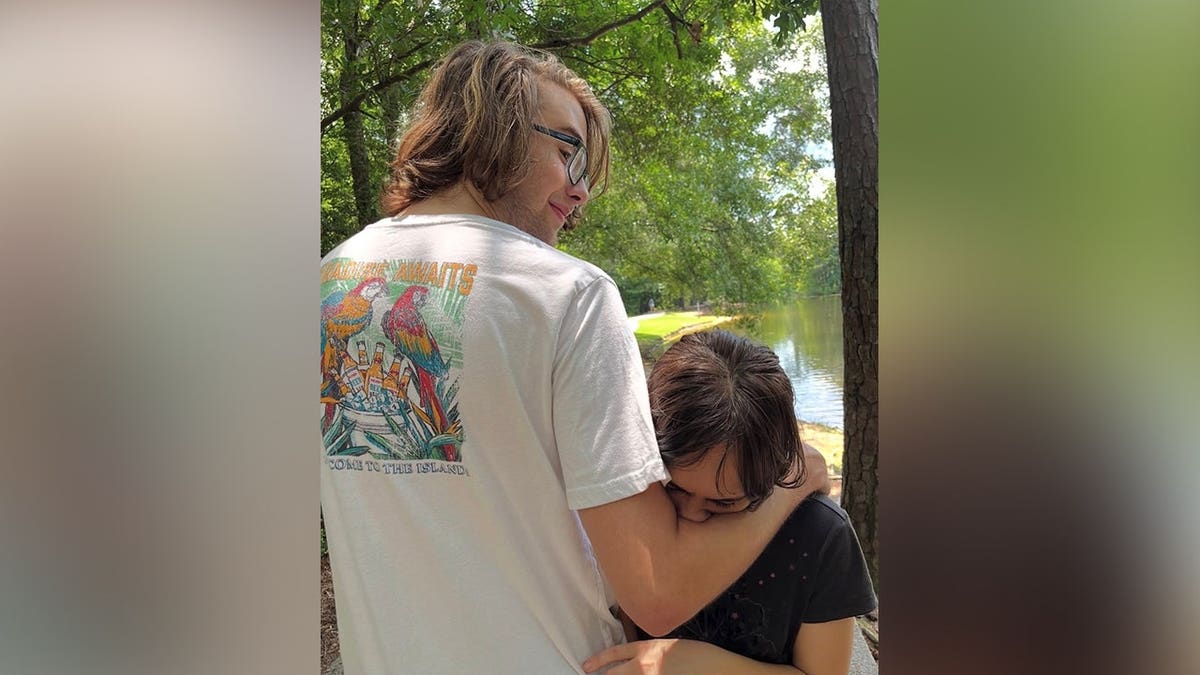 Julian Harris, wearing glasses and a white T-shirt, embraces Madison Duren beside a tree-lined body of water.