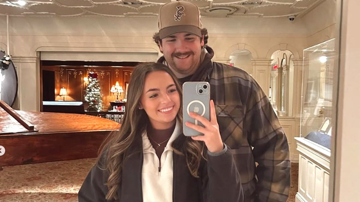 Leah Fonte smiles while taking a mirror selfie with her fiancé, firefighter Shane Hirstius, who stands behind her wearing a baseball cap and plaid jacket.