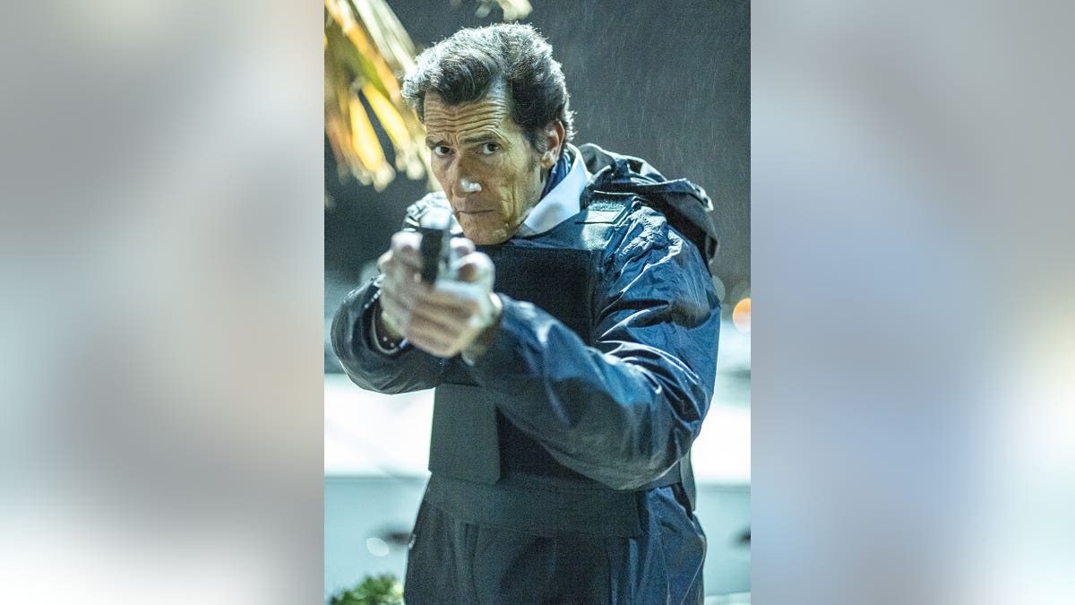 Geoffrey Moore wearing a jacket holding a gun and looking serious.