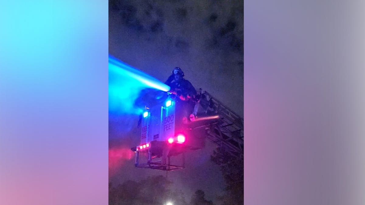 A firefighter operates a hose from an elevated fire truck ladder at night, spraying a stream of water illuminated by bright blue emergency lights.