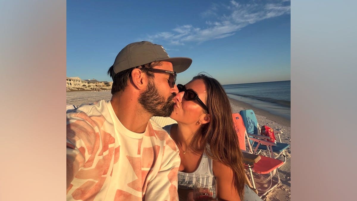 Jessie James Decker and husband Eric Decker share a kiss while sitting together on a sandy beach at sunset.