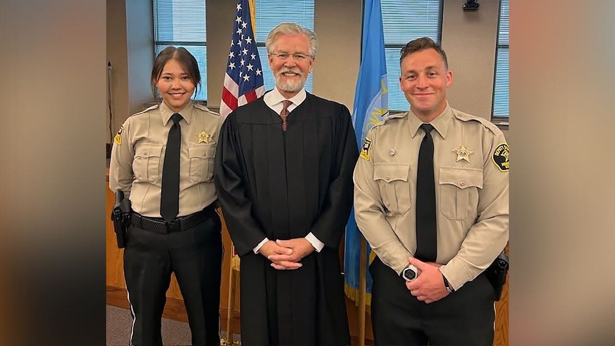 Deputy Katherine Frank stands beside a judge wearing a black robe and another uniformed sheriff's deputy, with American and South Dakota flags behind them.