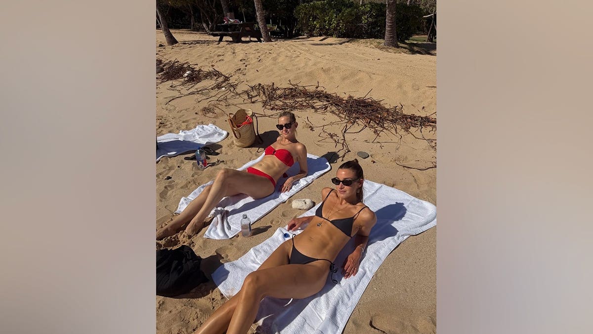 Kate Hudson relaxes in a red strapless bikini beside a friend wearing a black string bikini as they sunbathe on white towels on a sandy beach in Hawaii.