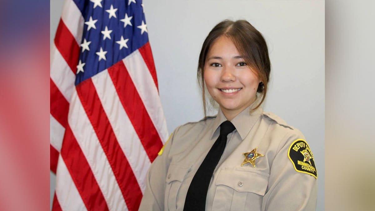 Minnehaha County Sheriff's Deputy Katherine Frank smiles in uniform beside an American flag, wearing a tan shirt, black tie and gold sheriff's badge.
