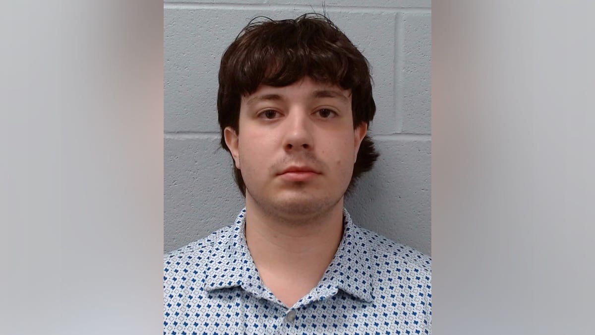 Booking photo of Noah Lara wearing a white button-down shirt with a blue geometric pattern against a gray cinder block wall.