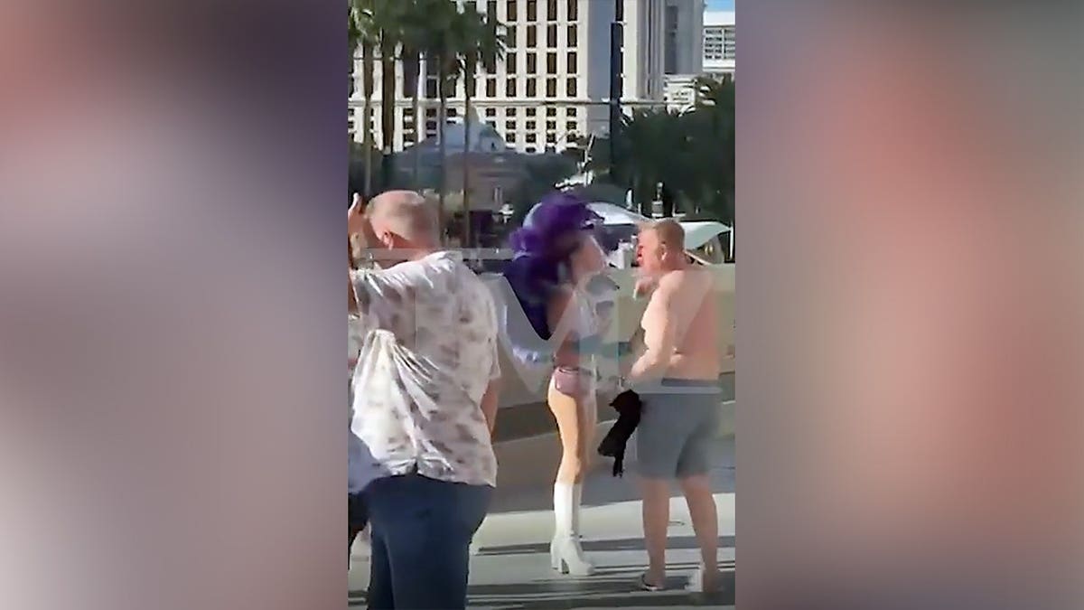 A shirtless man in gray shorts walks away from a woman wearing a white showgirl costume with large purple feathers near an escalator on the Las Vegas Strip.