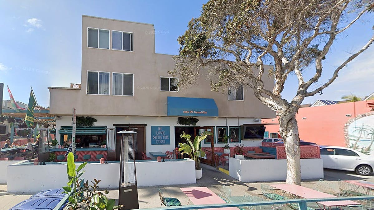 Street view of Miss B's Coconut Club in San Diego, showing a beige building with a blue awning, outdoor patio seating and a large tree near the entrance.