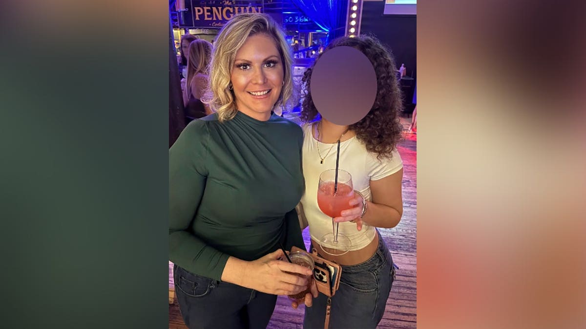 Misty Leslie, wearing a dark green top, smiles beside another person whose face is obscured as they hold drinks inside a brightly lit entertainment venue.