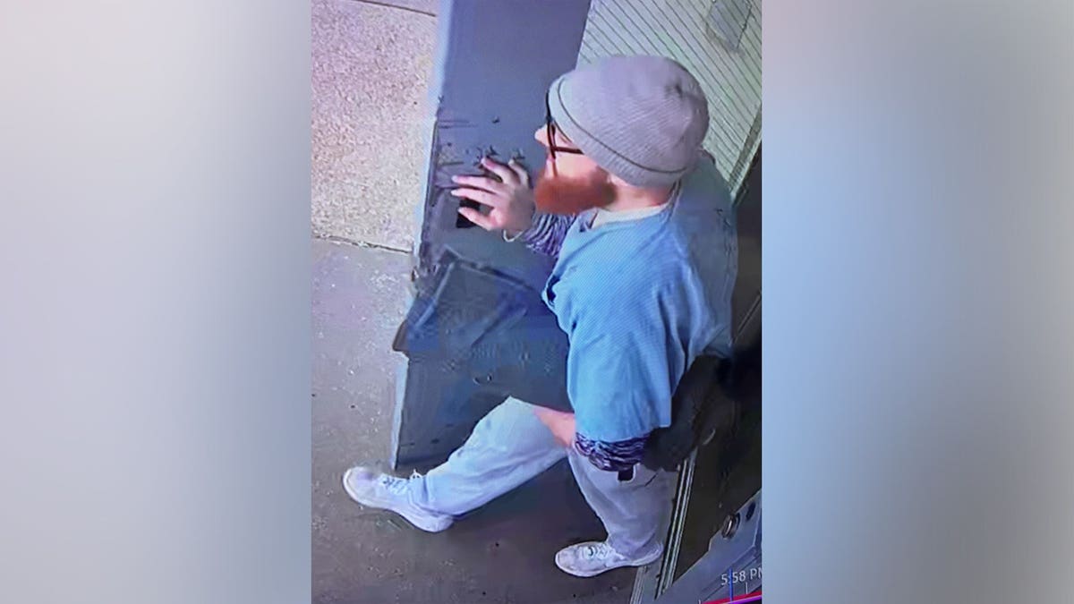 Overhead surveillance image of Jacob Allen Morley wearing a gray beanie, glasses, blue prison shirt, light-colored pants and white sneakers while walking through a prison facility.
