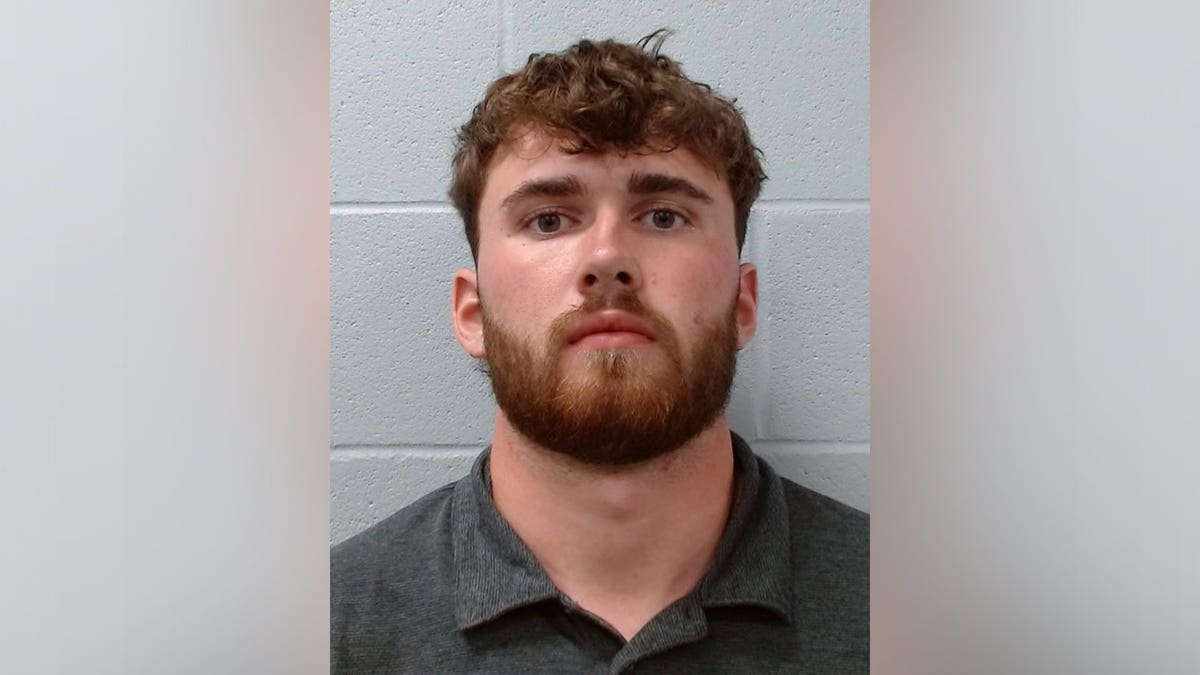 Booking photo of William Oldner, who has short brown hair and a beard, wearing a dark gray polo shirt against a gray cinder block wall.