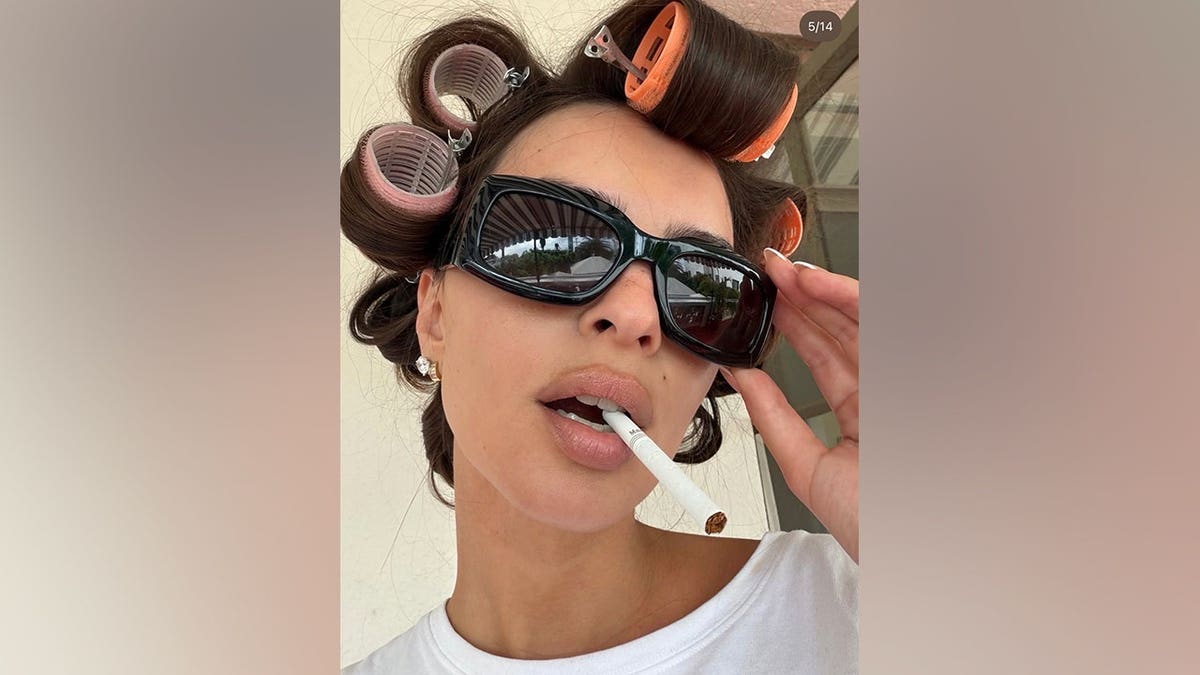 Emily Ratajkowski takes a close-up selfie wearing oversized sunglasses and colorful pink and orange hair rollers, with a cigarette between her lips.