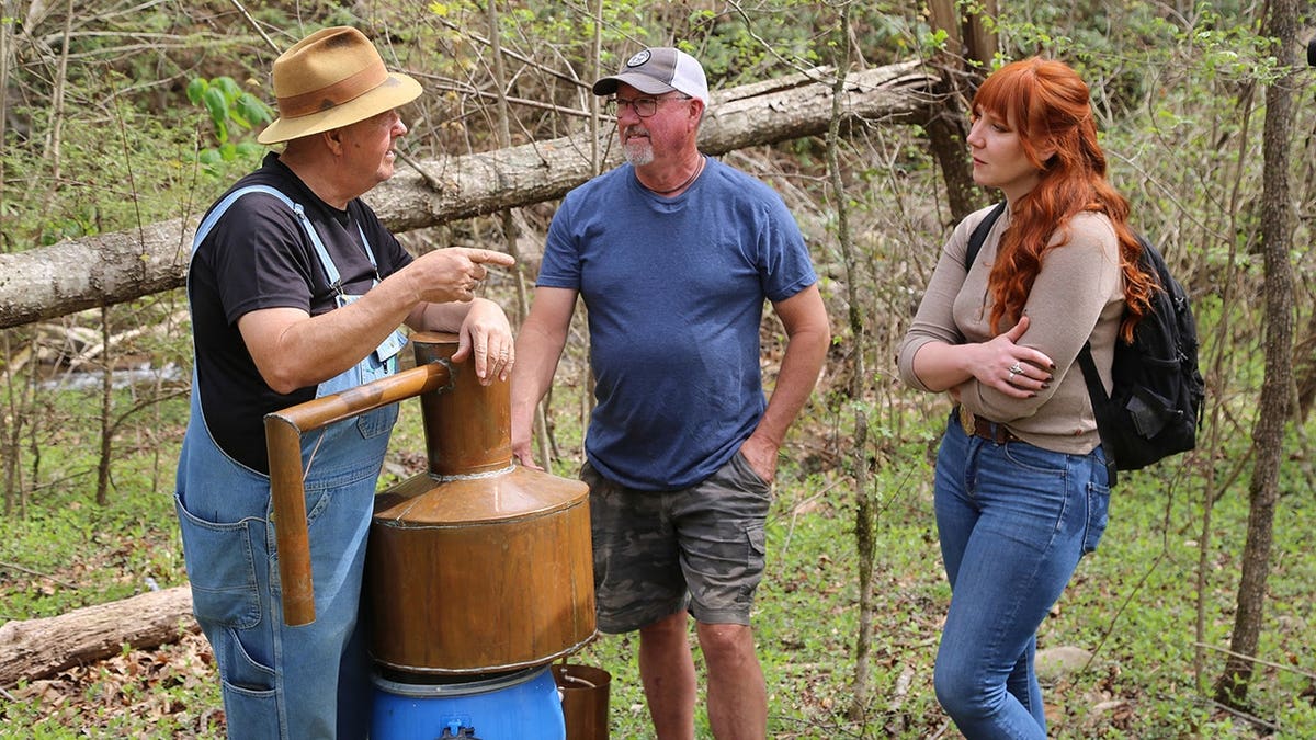 Amanda Bryant, Kelly Williamson and Mark Ramsey stand in woods near a mini still.