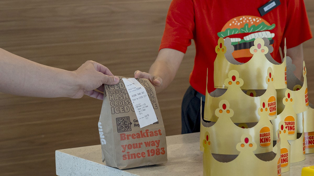 A Burger King employee hands a breakfast bag to a customer while standing behind paper crowns.