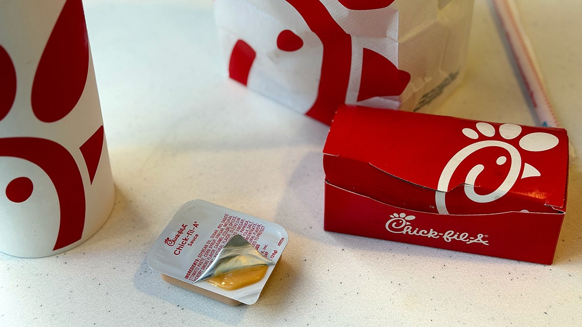 An opened pouch of Chick-fil-A Sauce next to a drink, bag and box of food.