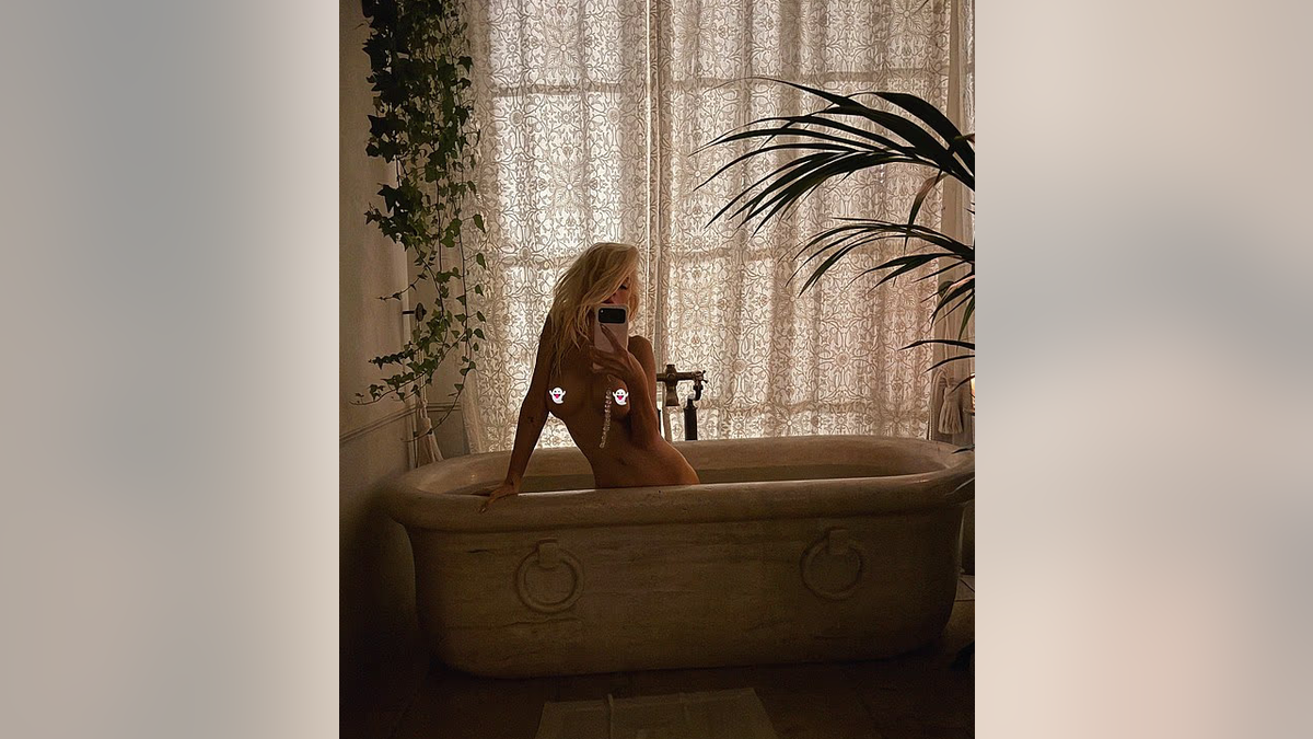 Christina Aguilera sits in a bathtub holding a smartphone in front of her face, with patterned curtains and plants behind her.