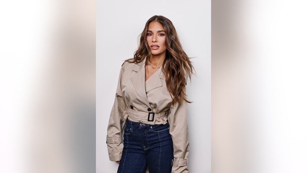 Jessie James Decker poses in a beige cropped jacket and dark blue jeans against a light gray background.