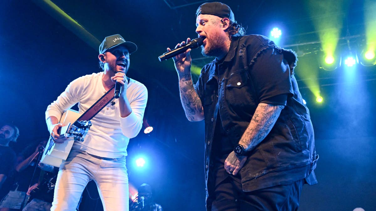 Drew Baldridge plays guitar and sings alongside Jelly Roll, who holds a microphone onstage under colorful lights.