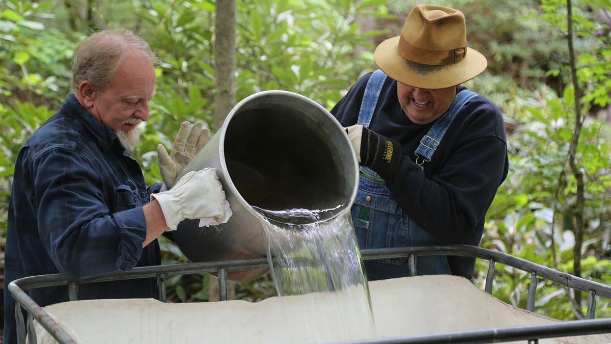 Eric Digger Manes and Mark Ramsey pour water into a tote in woods.