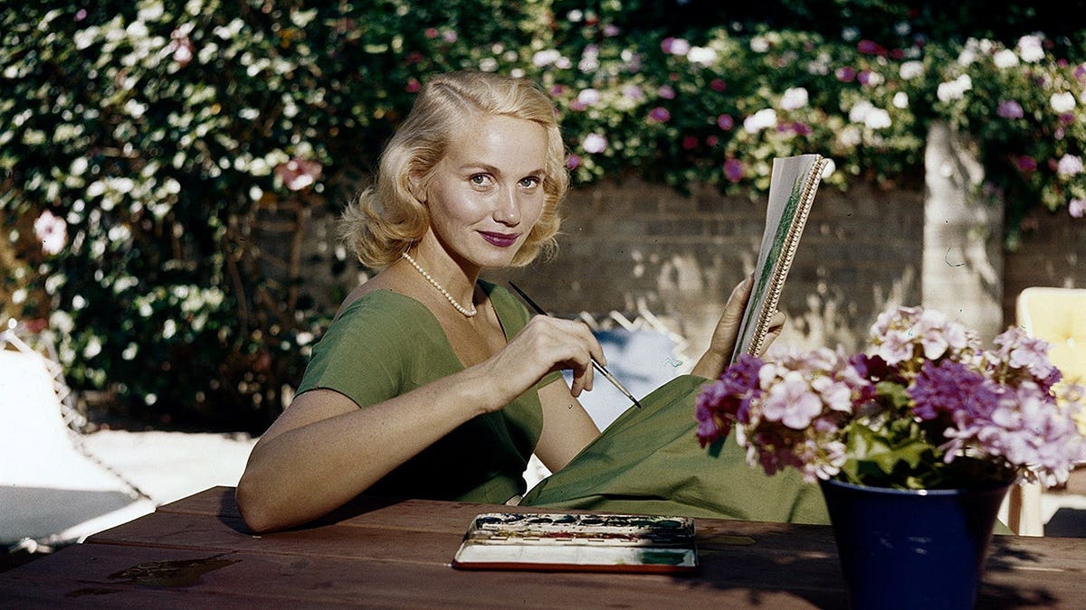 Eva Marie Saint paints in a garden, holding a sketchbook and paintbrush.