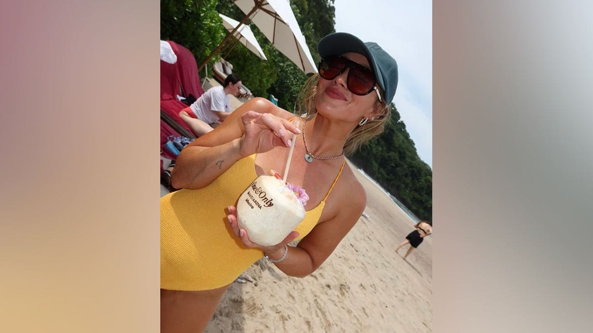 Hilary Duff poses on a sandy beach in a yellow one-piece swimsuit, holding a coconut topped with a flower while wearing sunglasses and a dark baseball cap.