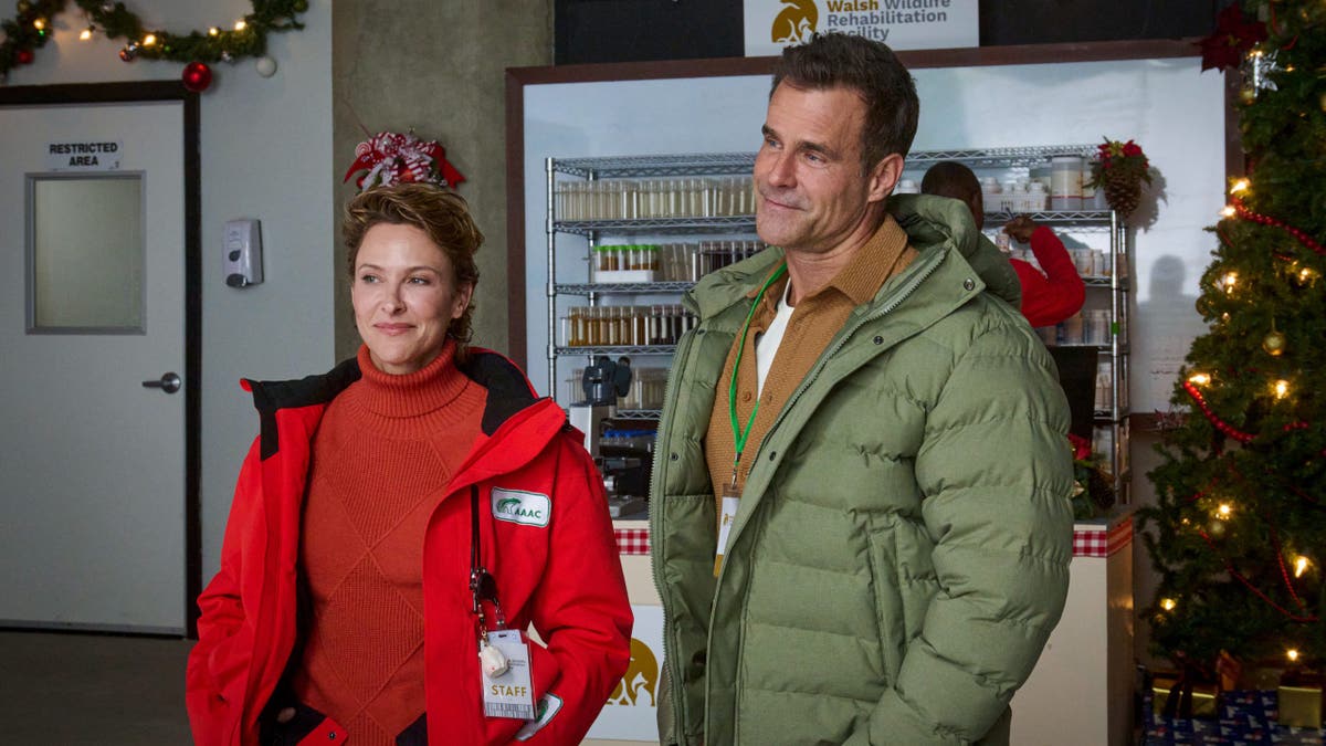 Jill Wagner stands beside Cameron Mathison inside a room decorated for Christmas, both wearing winter jackets.