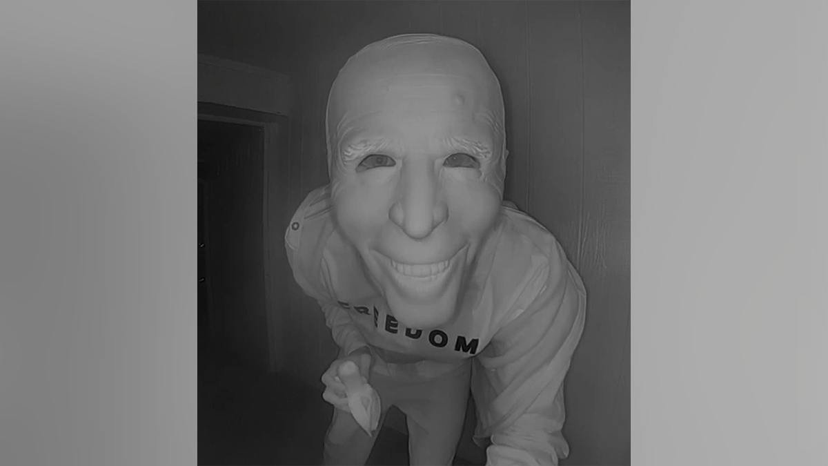 A close-up from doorbell footage shows a visitor leaning toward the camera in a grinning Joe Biden mask while holding a peeled banana