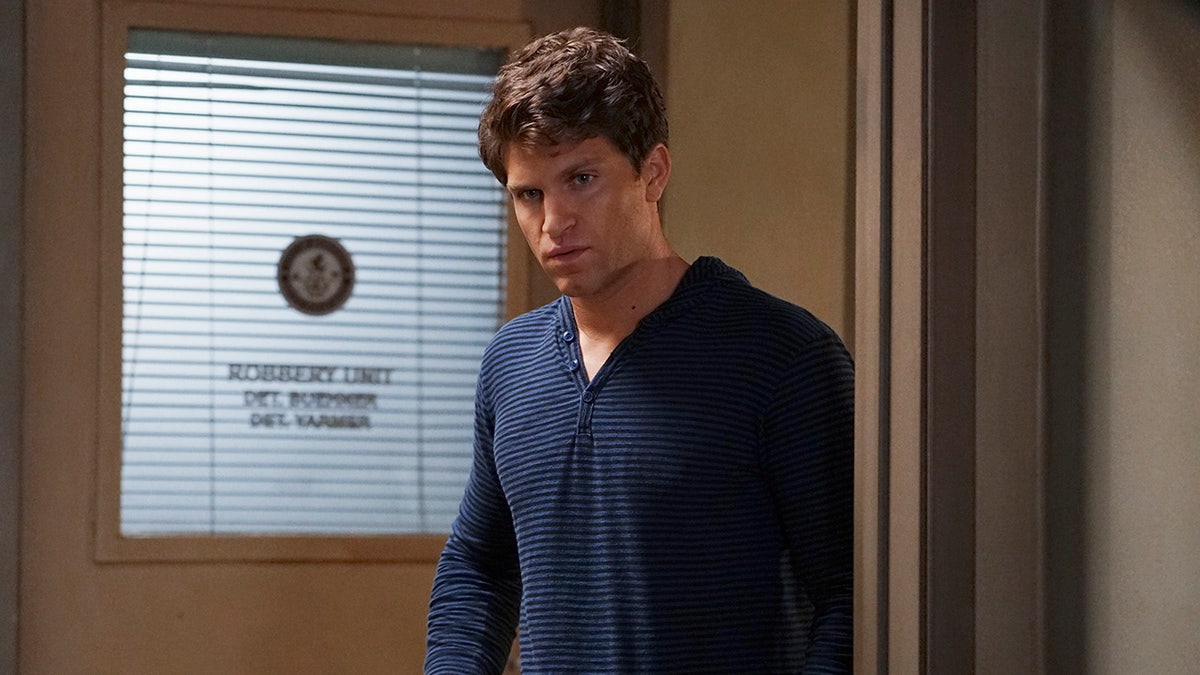 Keegan Allen wearing a dark blue striped shirt stands inside a room near a glass door with blinds.