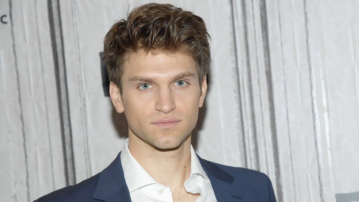 Keegan Allen wears a navy blue suit jacket and white shirt while facing the camera.