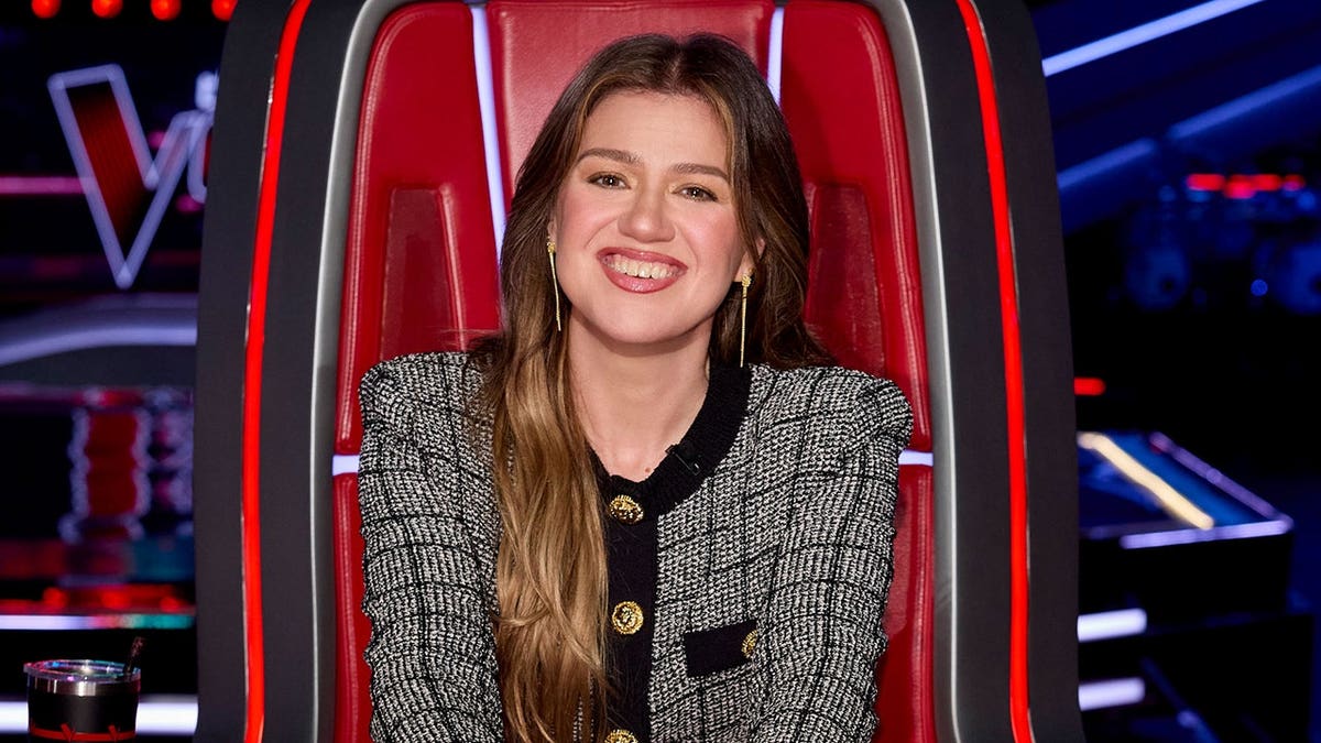 Kelly Clarkson sits in a chair on The Voice set.